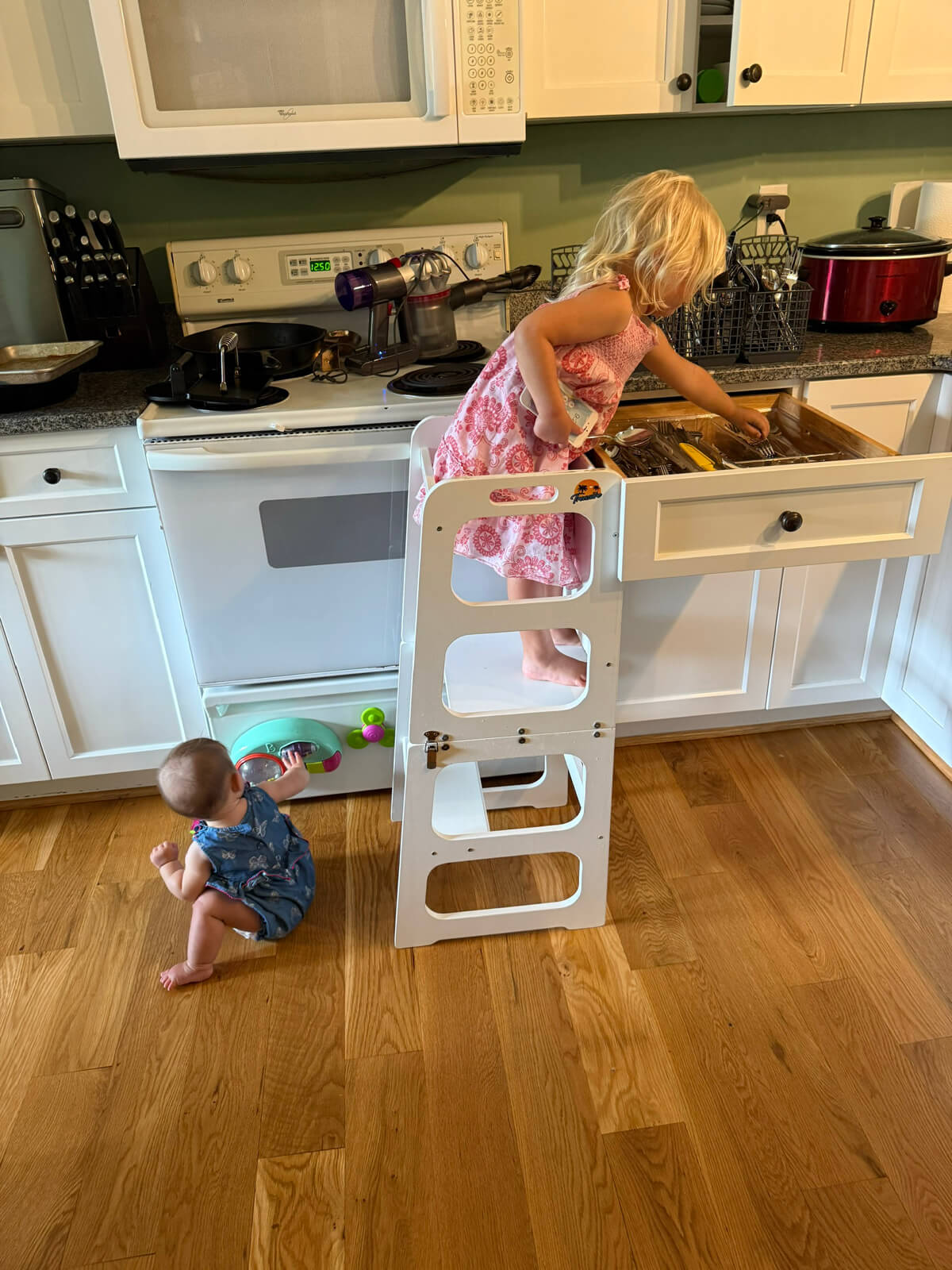 Toddler standing in learning tower helping unload silverware while baby crawls on floor. 