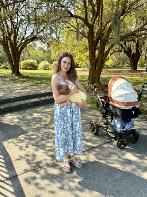 Mother standing in park in Charleston, SC, nursing newborn baby beside baby bassinet under live oak trees.