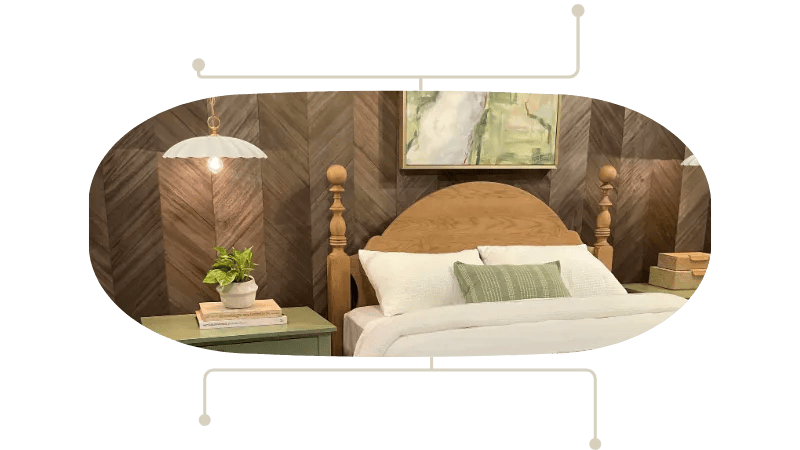 Cozy bedroom with a wooden headboard, white bedding, and a green throw pillow; a nightstand with a plant and books sits to the left, and a painting hangs above the bed.