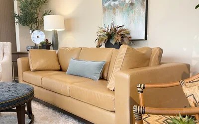 Tan leather sofa with a light blue cushion in a bright living room, with plants, a lamp, and an abstract painting on the wall.