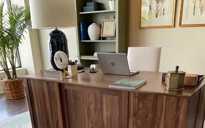 Wooden desk in a home office with a laptop, plant, lamp, bookshelf, and framed botanical art.