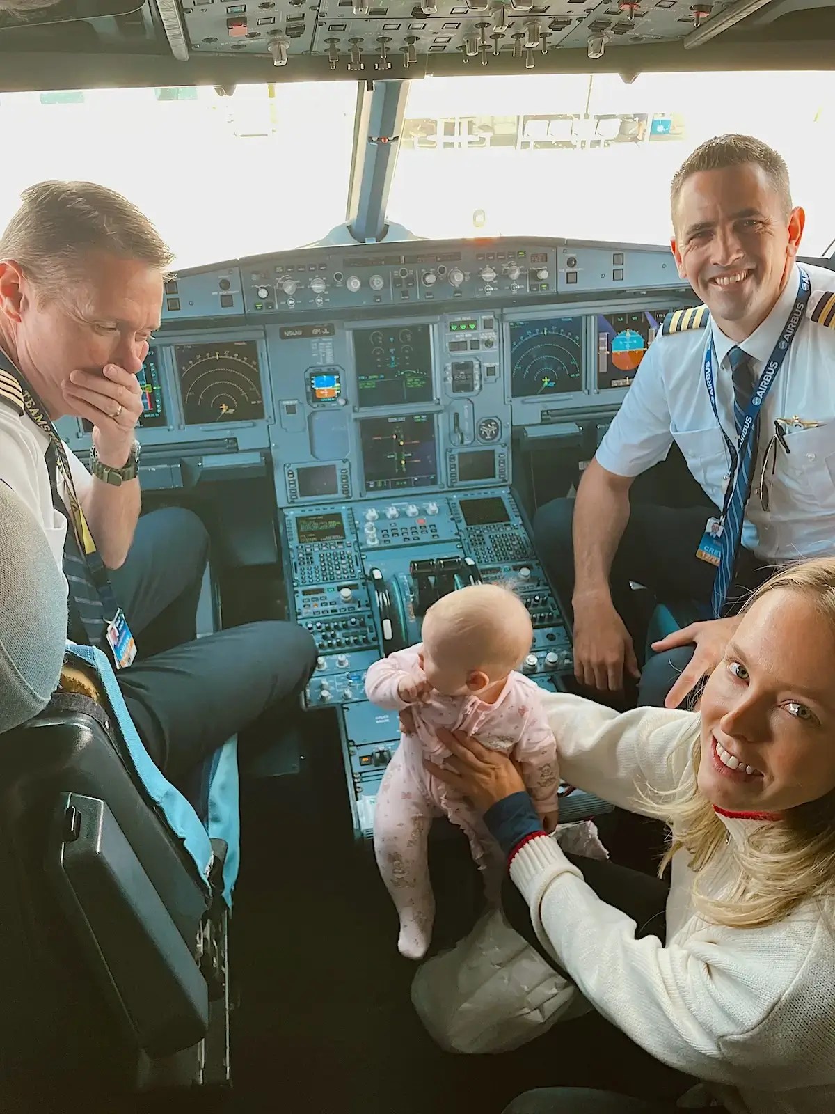 In the cockpit of an airplane with a baby and two pilots and her mom on the baby's first flight. 