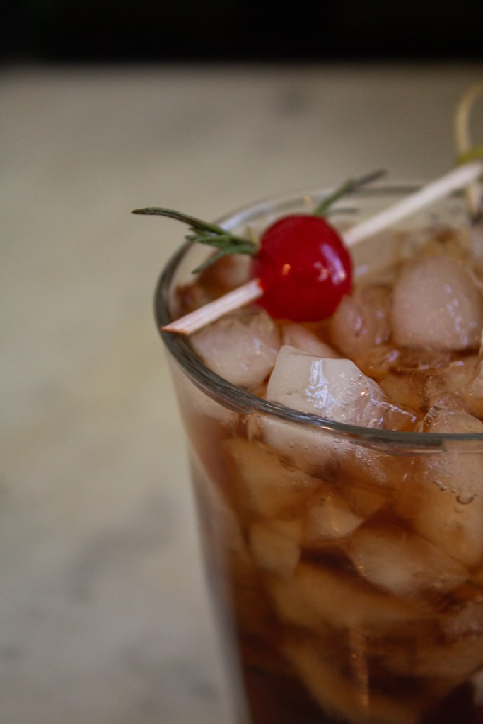Close-up of a drink with ice, garnished with a cherry and a sprig of herbs, set against a blurred background.