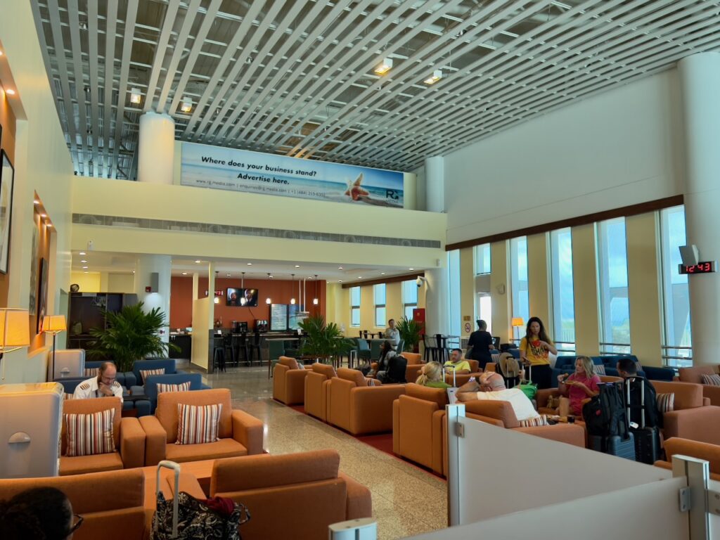 Travelers relax in a spacious airport lounge with orange armchairs, a bar, and tall windows