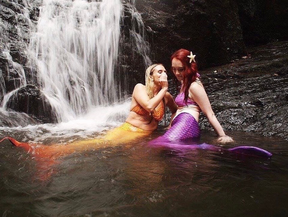 It Was All a Dream photo by artist Diane Landro featuring two people dressed as mermaids, one with a golden tail and the other with a purple tail, sit at the base of a waterfall.