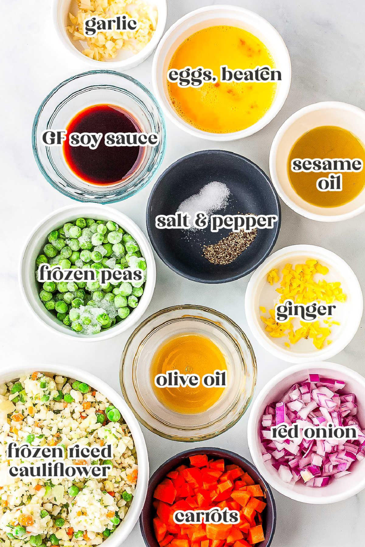 A flat lay of labeled ingredients in small bowls needed to make cauliflower fried rice, including minced garlic, beaten eggs, gluten-free soy sauce, frozen peas, salt and pepper, sesame oil, minced ginger, olive oil, frozen riced cauliflower, diced red onion, and diced carrots.
