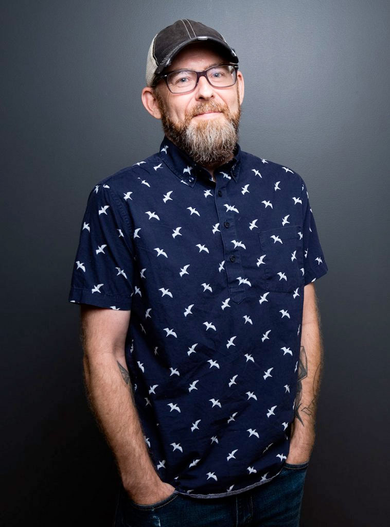 A bearded man in glasses and a cap stands against a plain dark background, wearing a navy blue shirt with a white bird pattern and his hands in his pockets.