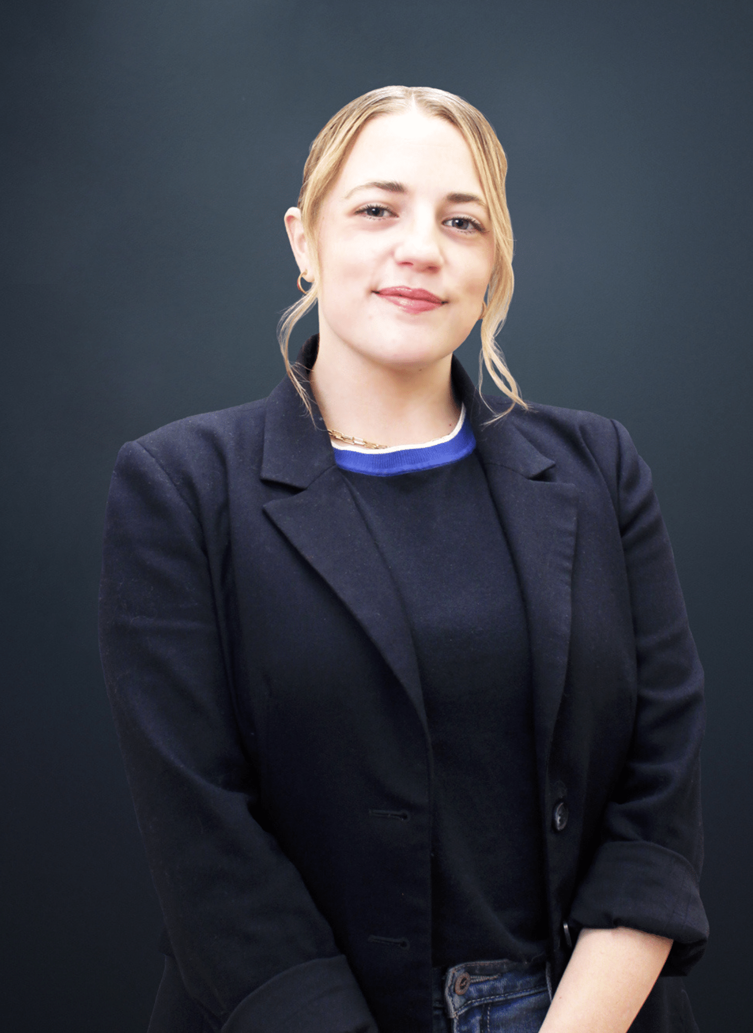 A person with light skin and blonde hair wearing a black blazer and dark sweater, standing in front of a plain dark background, smiling slightly at the camera.