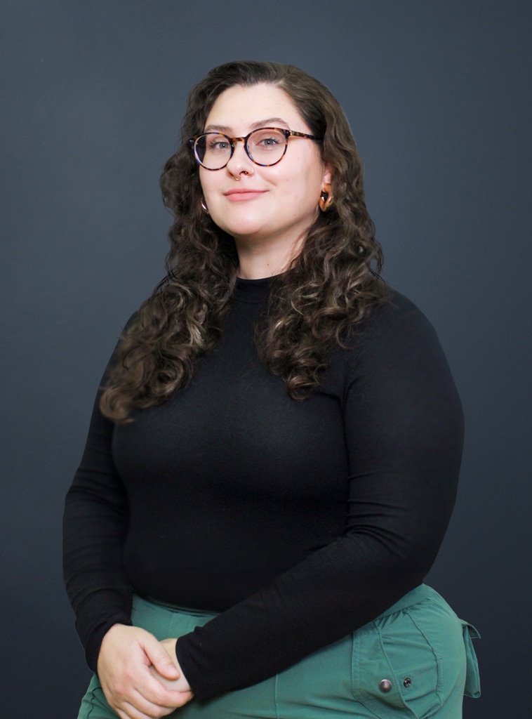 A person with long curly brown hair, wearing glasses, a black long-sleeve top, and green pants, stands against a plain dark background, smiling slightly with hands folded.