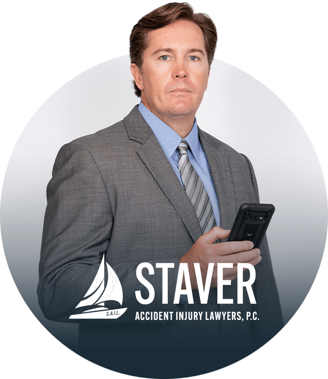 A man in a gray suit holding a smartphone looks at the camera; the logo and text Staver Accident Injury Lawyers, P.C. appear at the bottom of the image.
