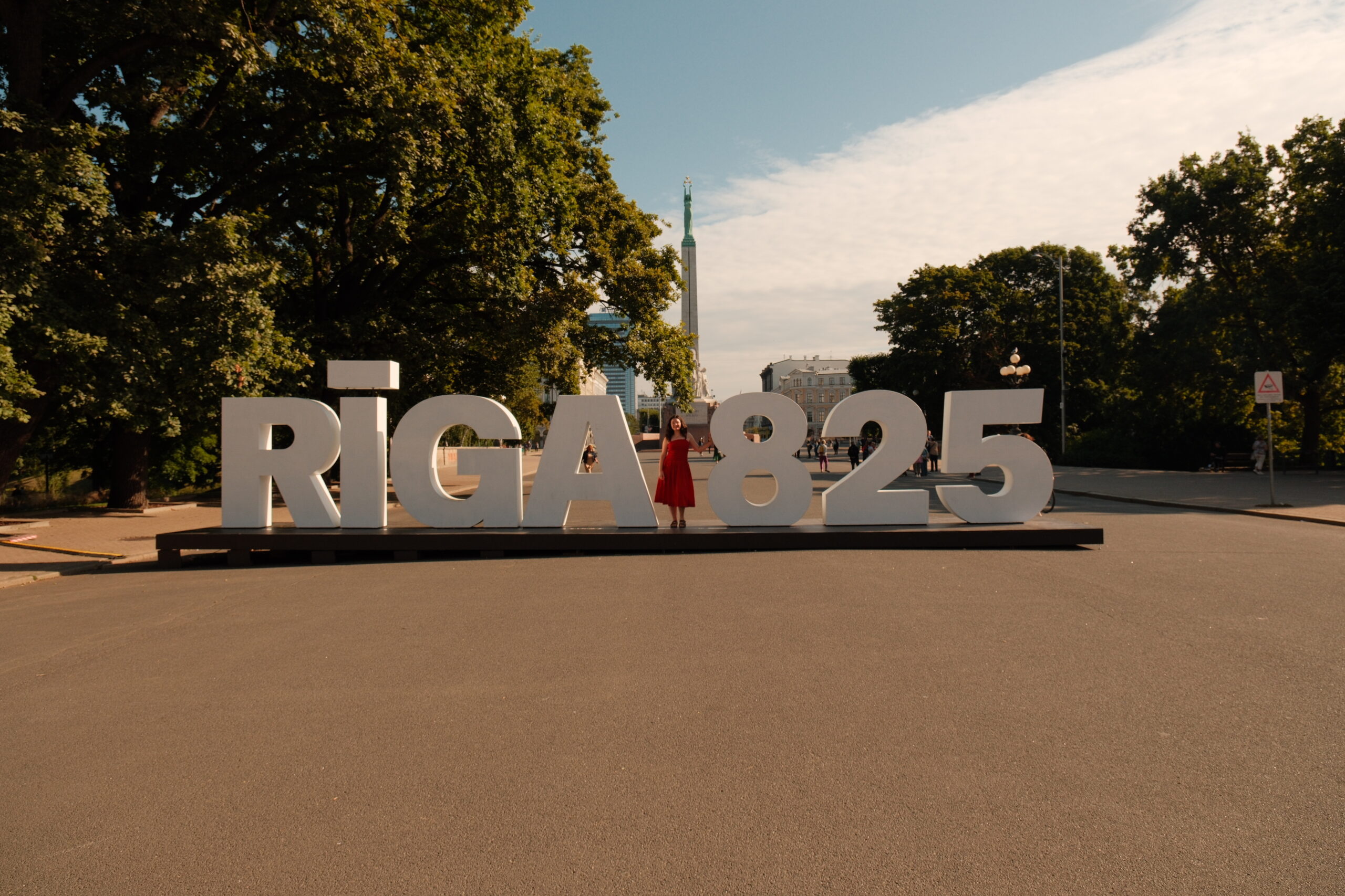 A woman in a red dress stands between oversized white letters spelling 'RIGA825' in a sunny plaza with trees in the background.