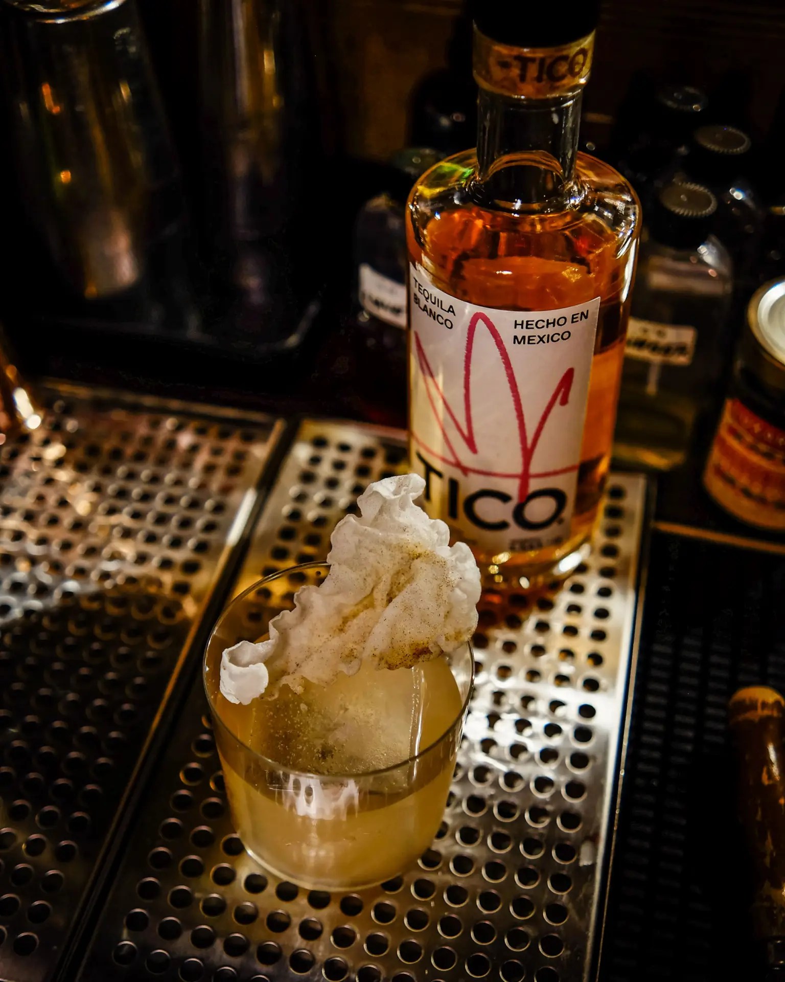 A finished cocktail sits in a rocks glass with block ice and a fried rice paper garnish, with a bottle of Tico Tequila behind it to the right