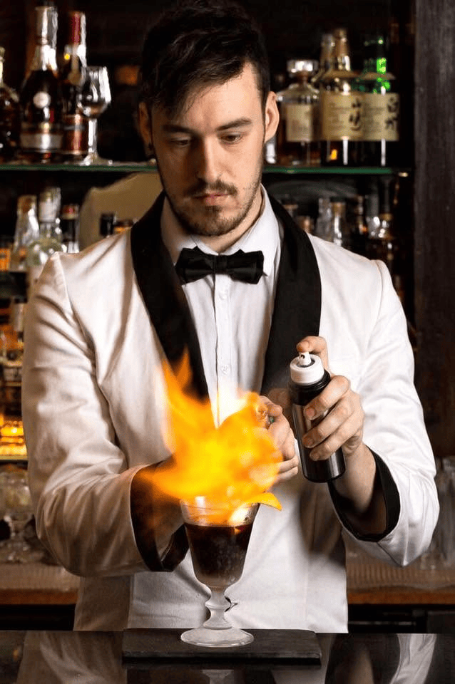 Bartender in a white tuxedo flaming a cocktail behind a hotel bar