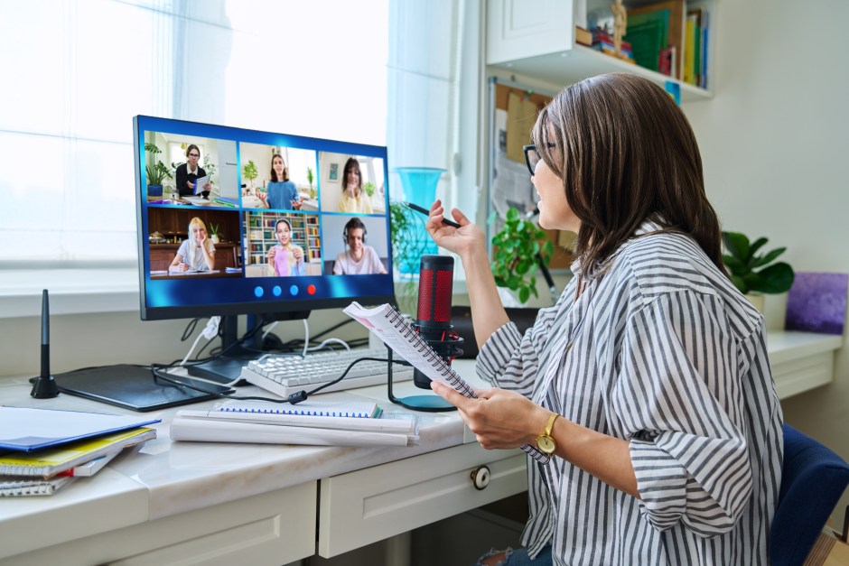 Female educator conducting an online lesson from home, holding a notebook and pen while engaging with students on a video call displayed on a computer monitor; educational materials and a microphone are on the desk