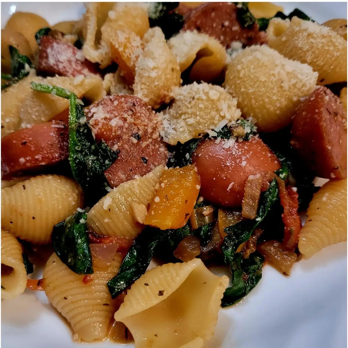 Sausage, vegetables, and shells are on a white plate garnished with parmesan cheese.