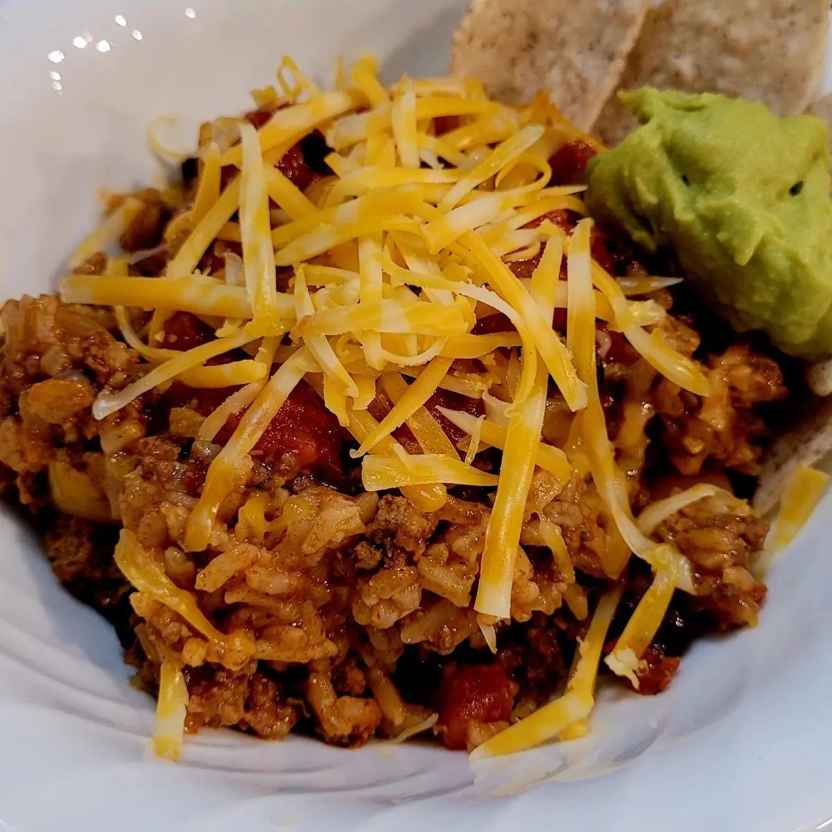 Stuffed pepper casserole made from rice, beef, and peppers is topped with shredded cheese, guacamole, and served with a side of tortilla chips in a white bowl.