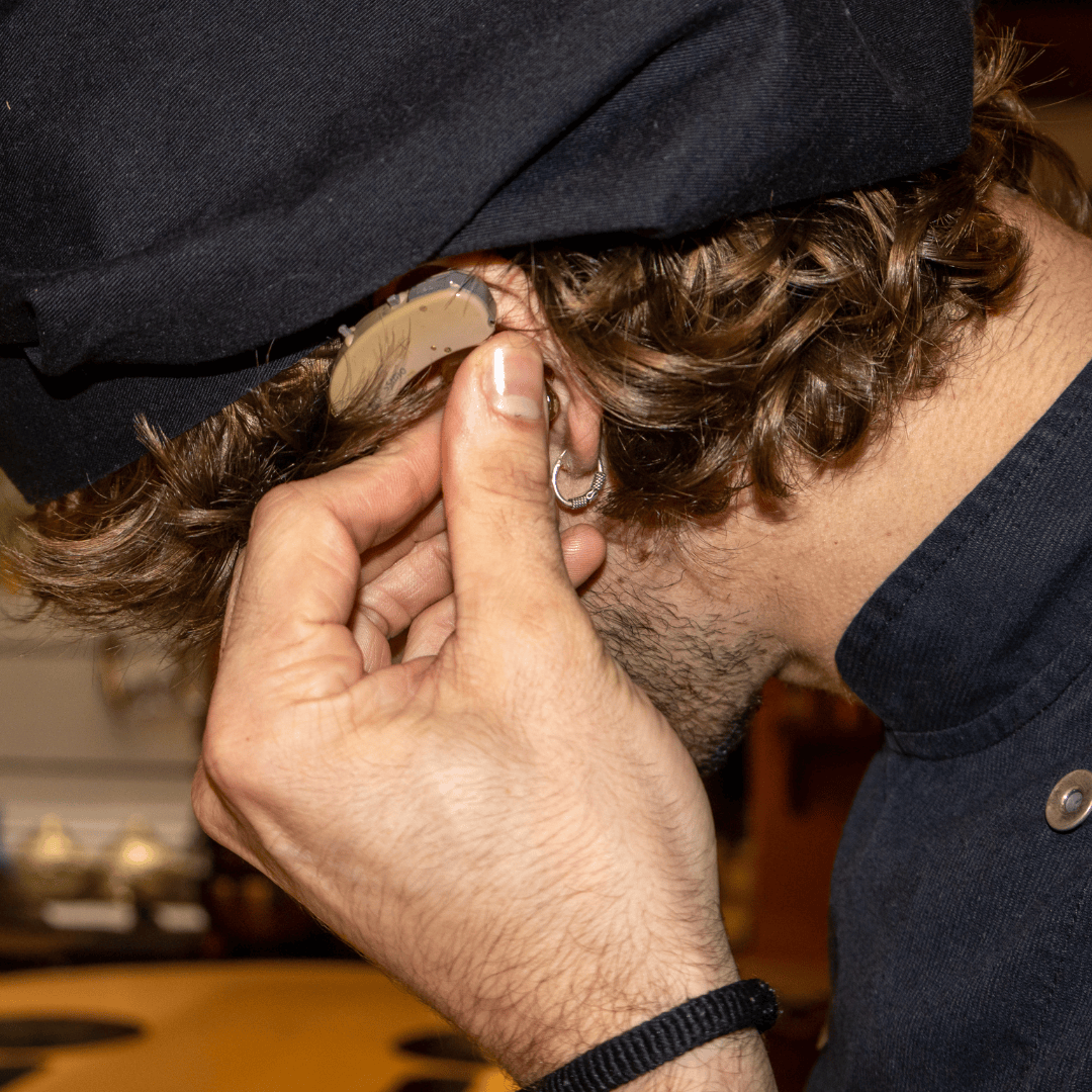 A person with curly hair adjusts their hearing aid behind their ear, wearing a black hat and dark clothing.