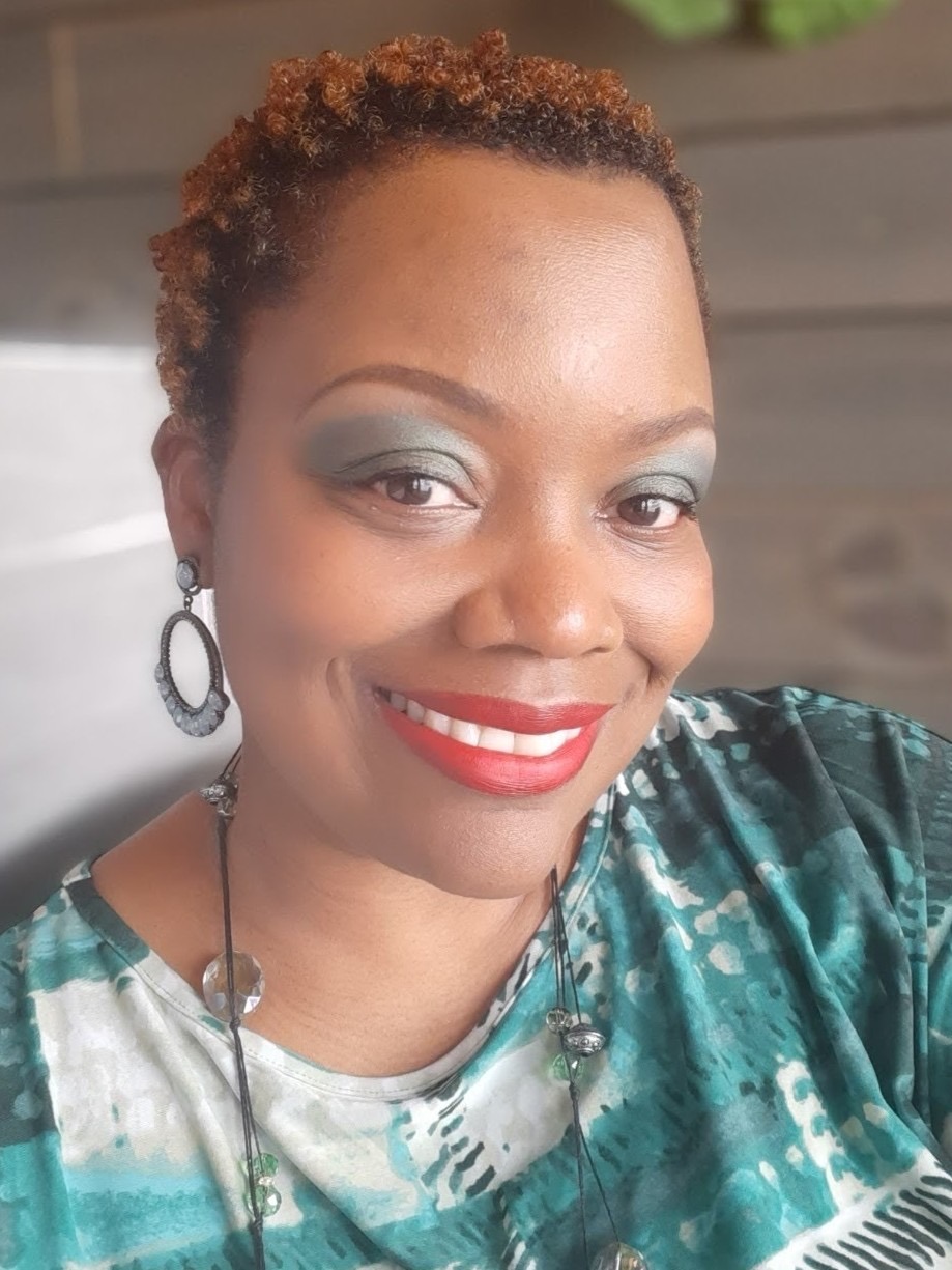 Woman with short curly hair and bold makeup smiles warmly. She wears colorful clothing and statement earrings, set against a soft-focus background.