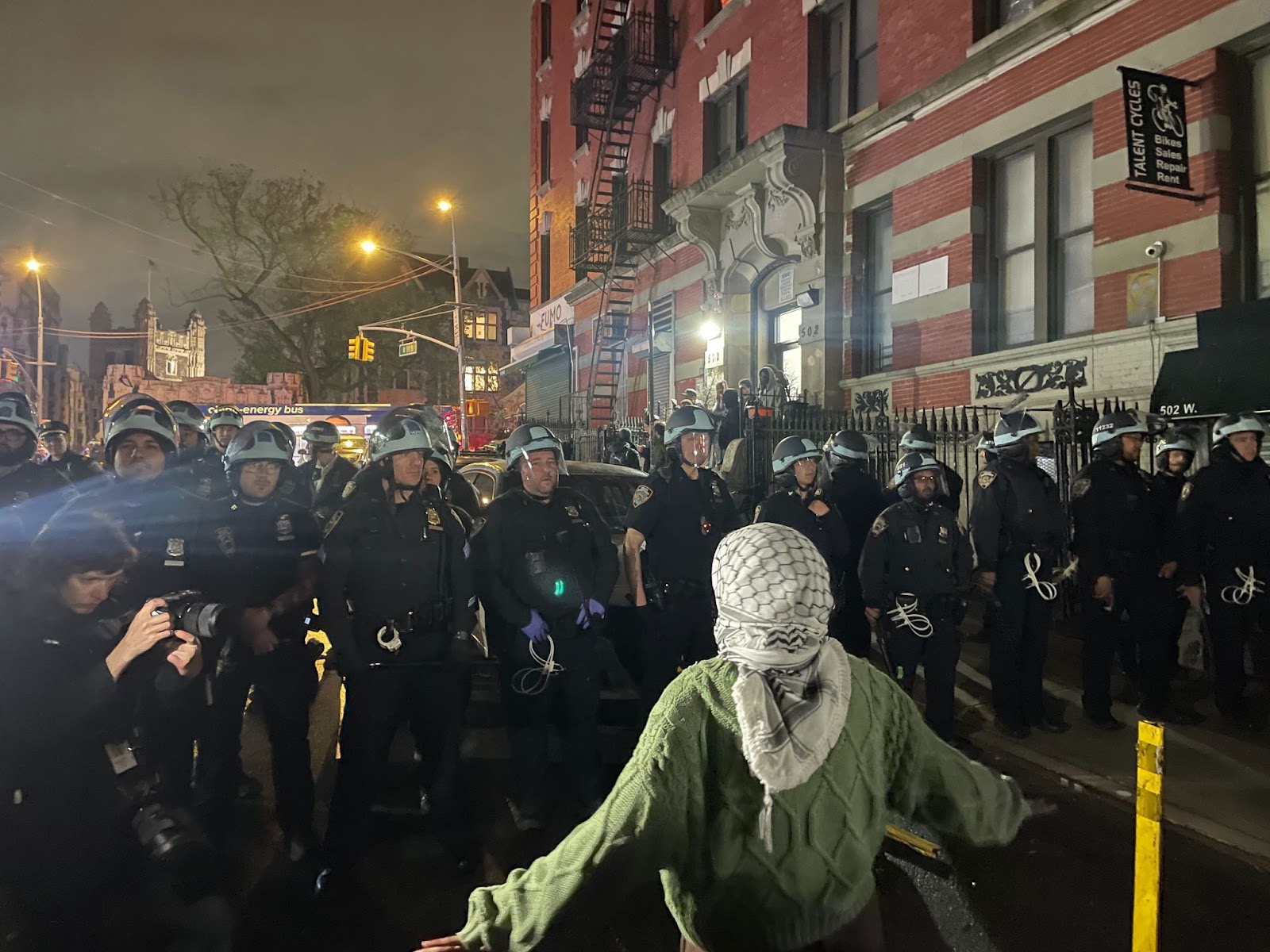 color photograph of a person wearing a white and black keffiyah on their head standing with their back to the camera facing a wall of militarized police