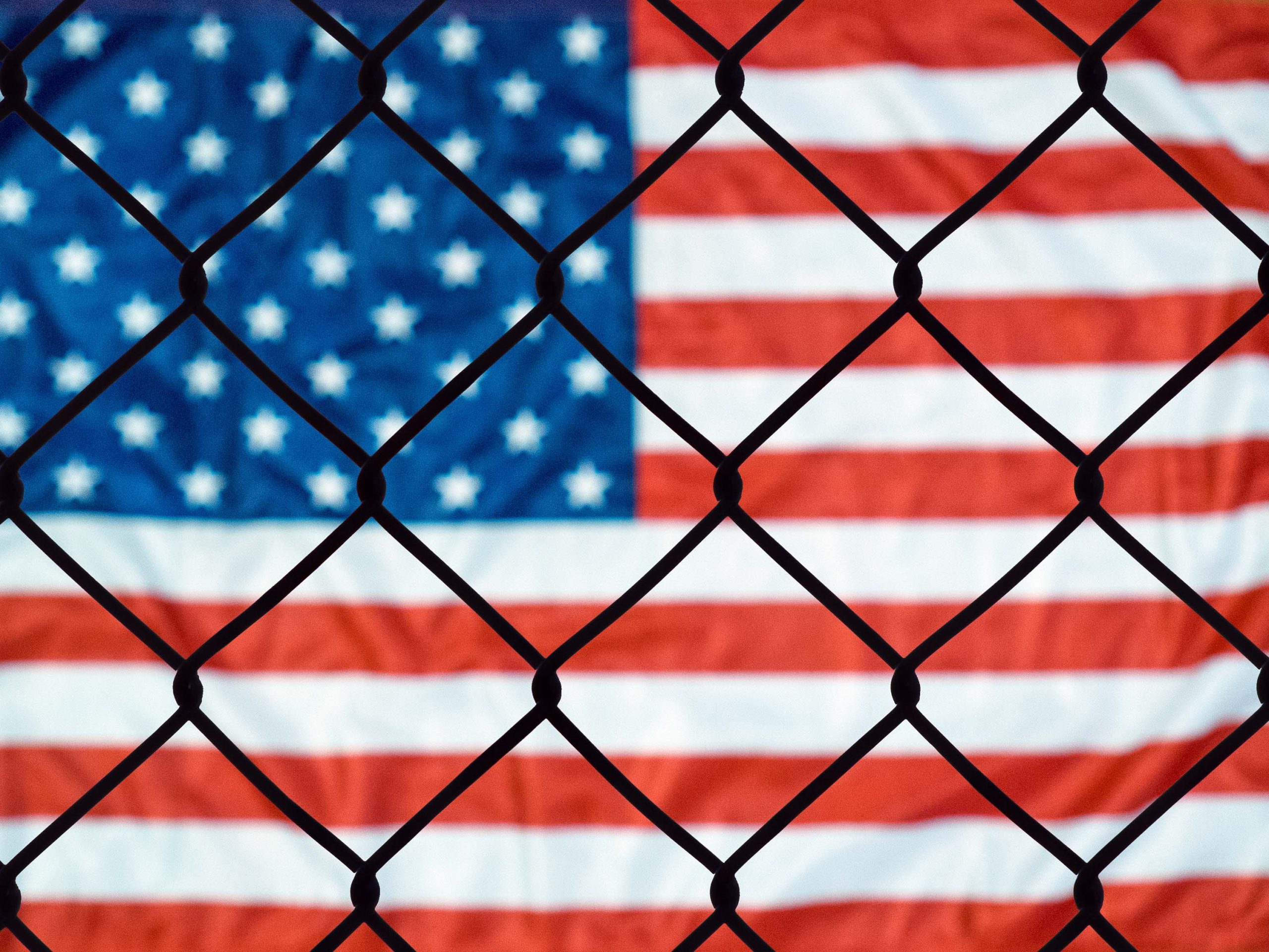 A stock photo shows an American flag is seen through a chain link fence.