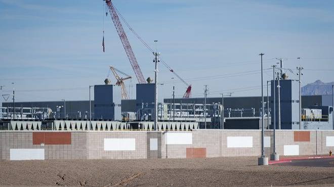 An industrial construction site with cranes rising behind a perimeter wall.