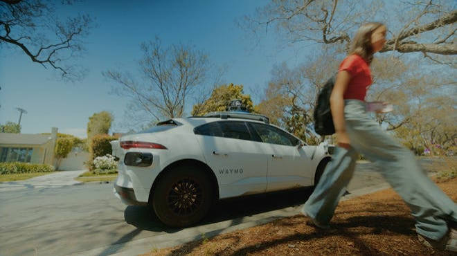 A white Waymo self-driving car parked on a street as a pedestrian walks past.