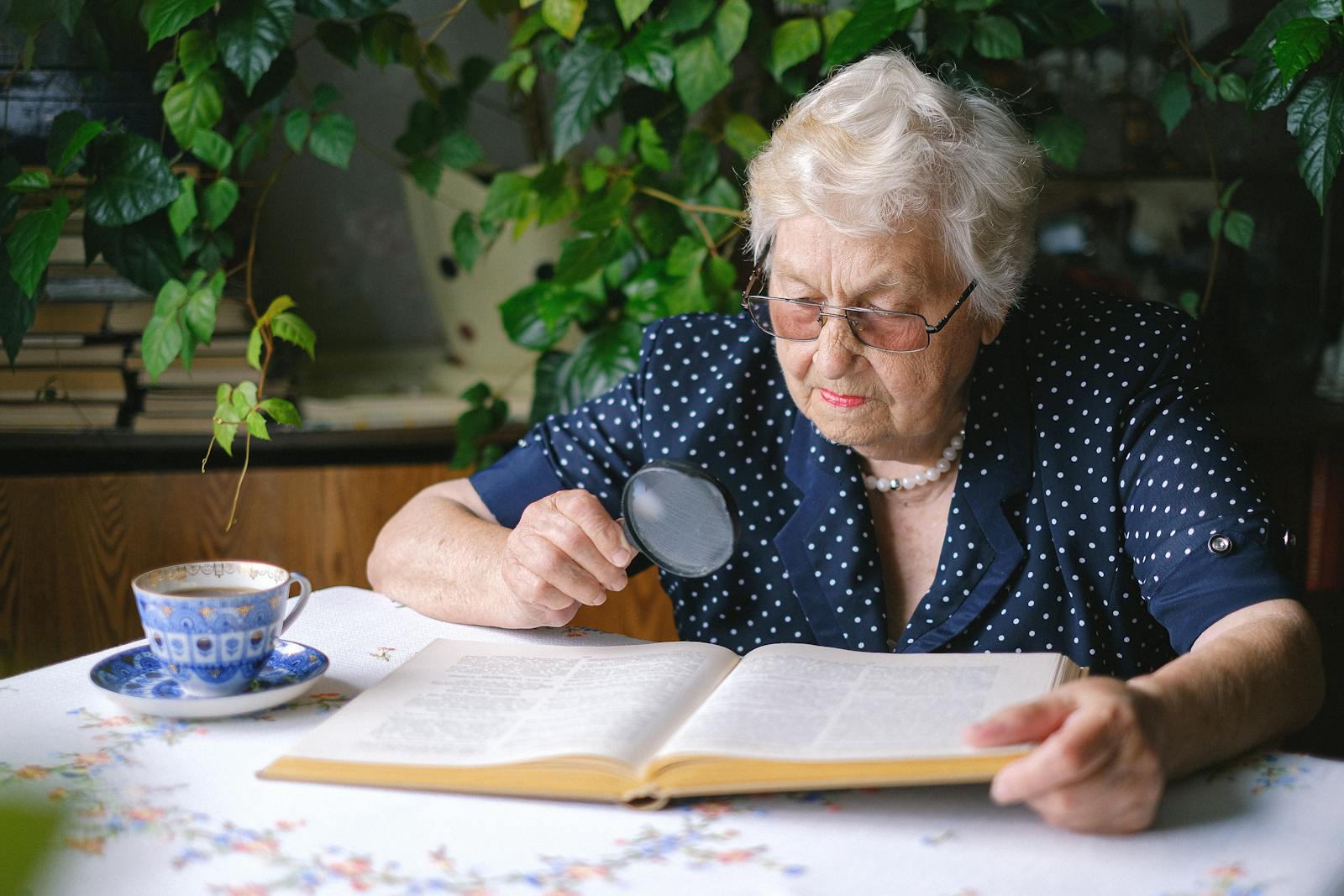 Une femme âgée lit un livre à la maison avec une loupe, entourée de plantes.