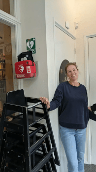 Vrouw staat binnen naast een wandgemonteerde Philips AED in houder met AED-bord erboven.