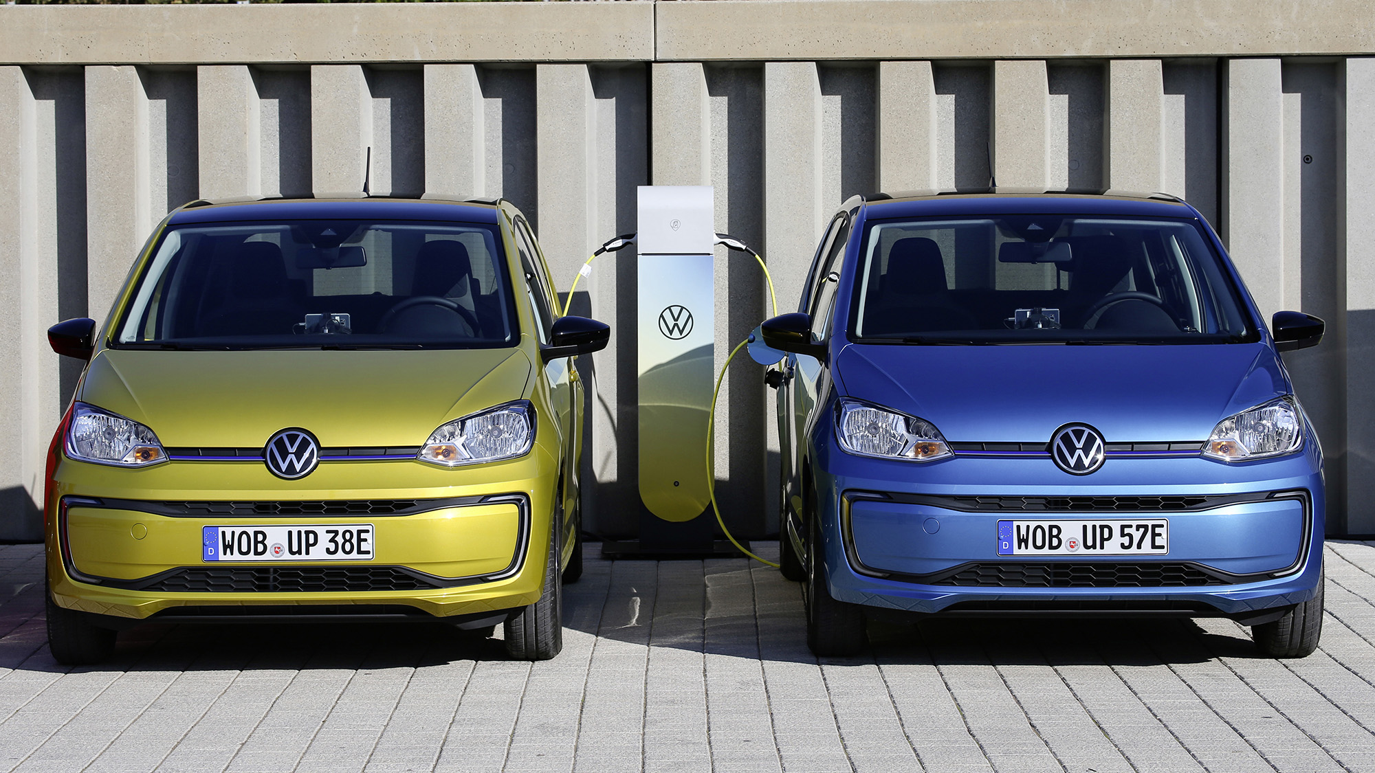 Dois VW e-Up!, um azul e outro amarelo carregando vistos de frente