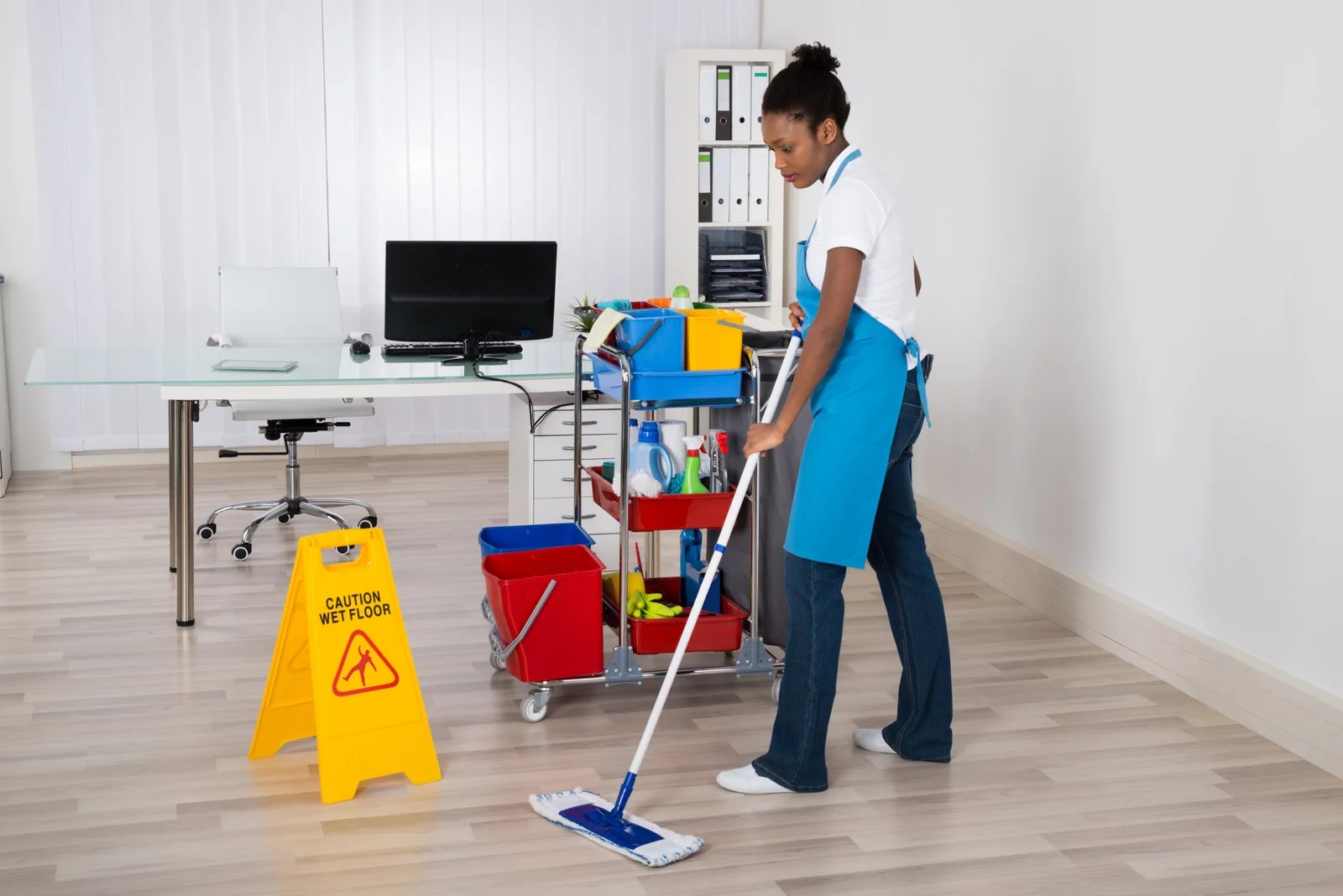 Female Janitor Mopping Wooden Floor With Caution Sign In Office