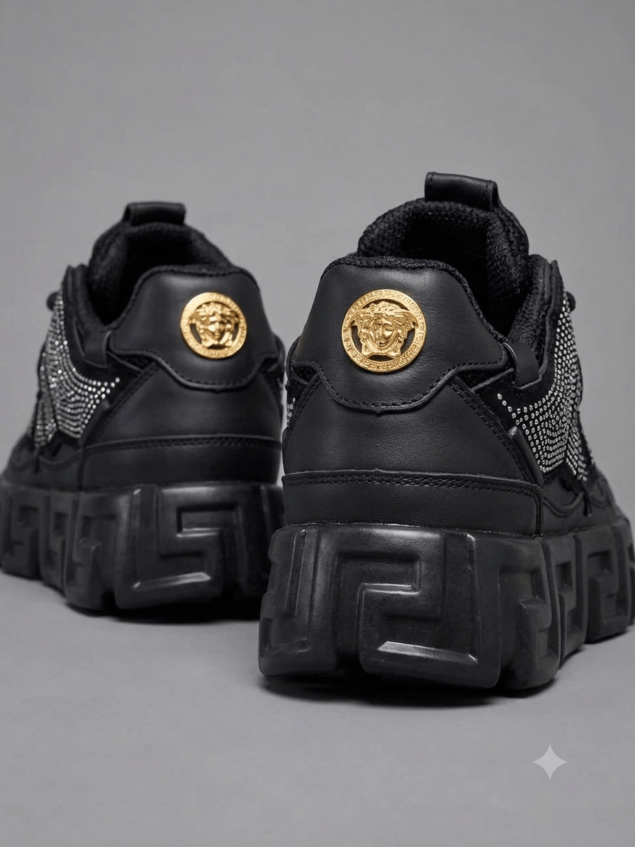 A pair of black Versace Chain Reaction platform sneakers decorated with all-over black rhinestones and a gold Medusa head on the tongue, floating over a plain gray background.