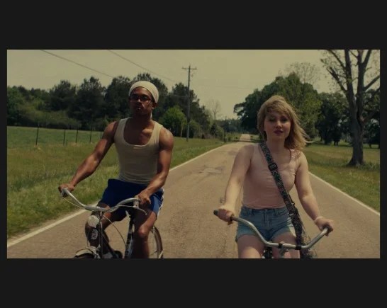 two adolescent siblings ride bicycles on a country road. The girl is white with blonde hair. She wears a pink sleeveless shirt and jean shorts The boy is black and wears a grey stocking cap, jean shorts and a white tank top