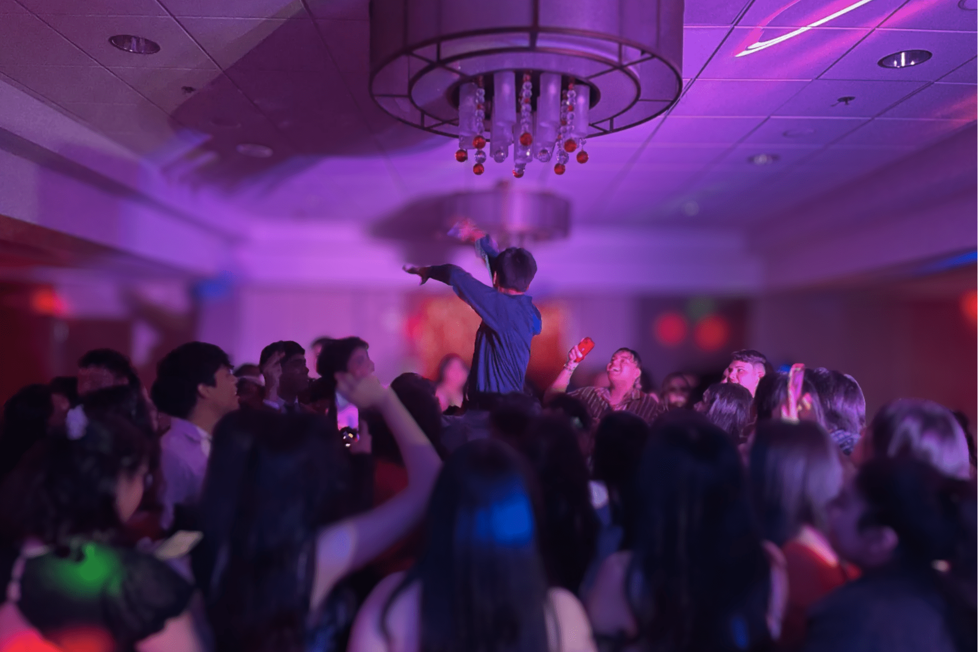 Crowd dancing under colorful lights at a packed party with the DJ hyping up guests on the dance floor.