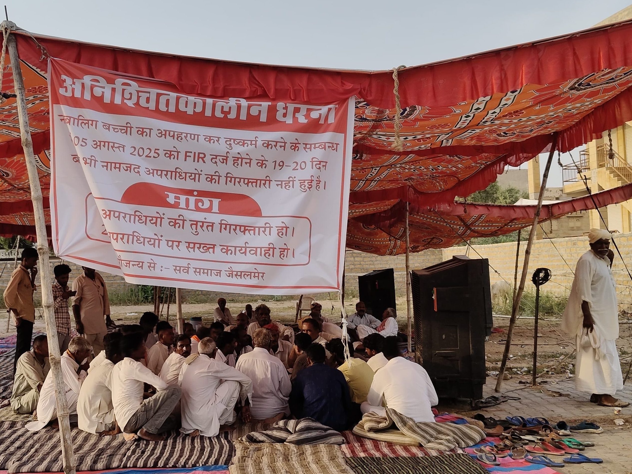 People seated beneath a canopy at a gathering with a Hindi banner.