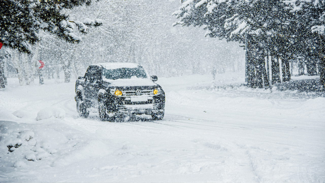 Extreme cold preparedness. SUV driving through heavy snowfall on a snow-covered road surrounded by trees. Learn how to stay warm during winter weather.