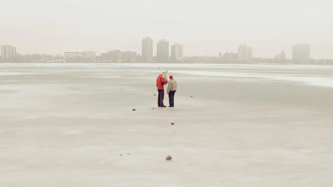 A couple standing on a vast, empty landscape with a city skyline blurred in the background, creating a serene and contemplative atmosphere.