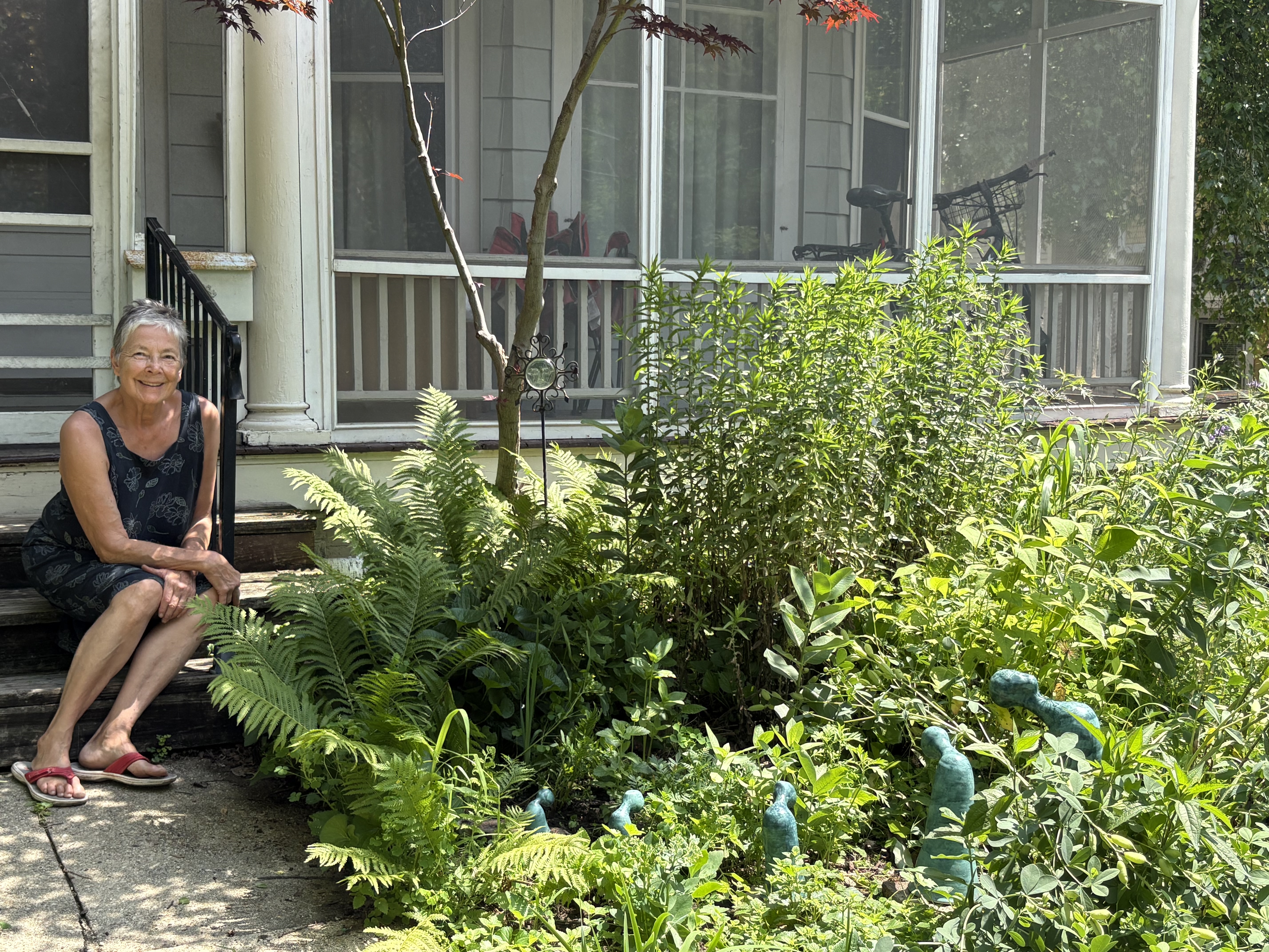 Woman with white hair sitting on porch steps near greenery and teal sculptures.