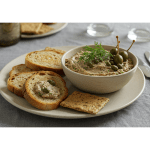 A stylishly plated bowl of mackerel pâté, garnished with fresh dill and capers, accompanied by an assortment of crispy bread and crackers, set on a cozy dining table with soft lighting
