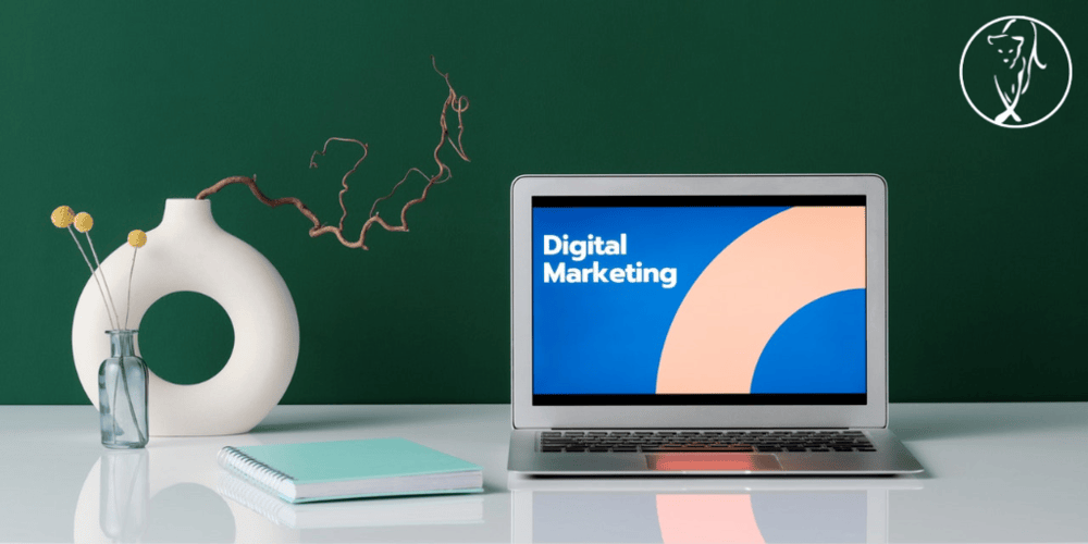 Laptop on a desk showing a Digital Marketing presentation slide, beside a vase and notebook against a green wall