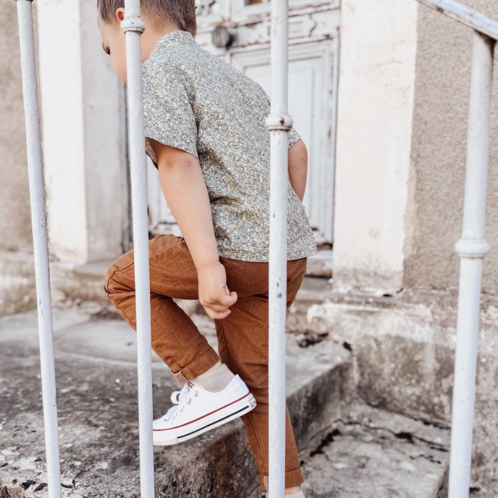 Un petit garçon de 3 ans vêtu d'une chemise col Mao fleuri et d'un pantalon en lin lavé, montant des escaliers en pierres.