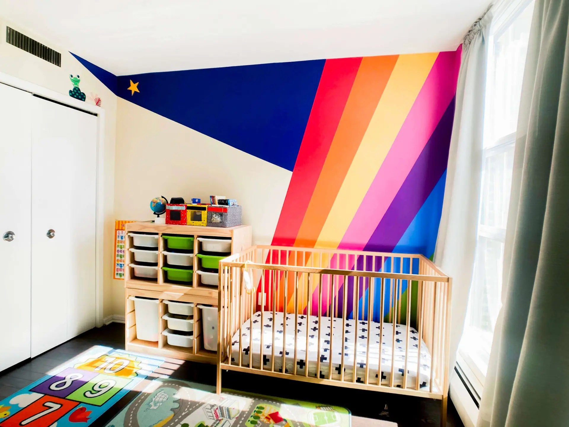 Nursery showing a IKEA crib and a Trofast toy storage with a big rainbow mural in the background.