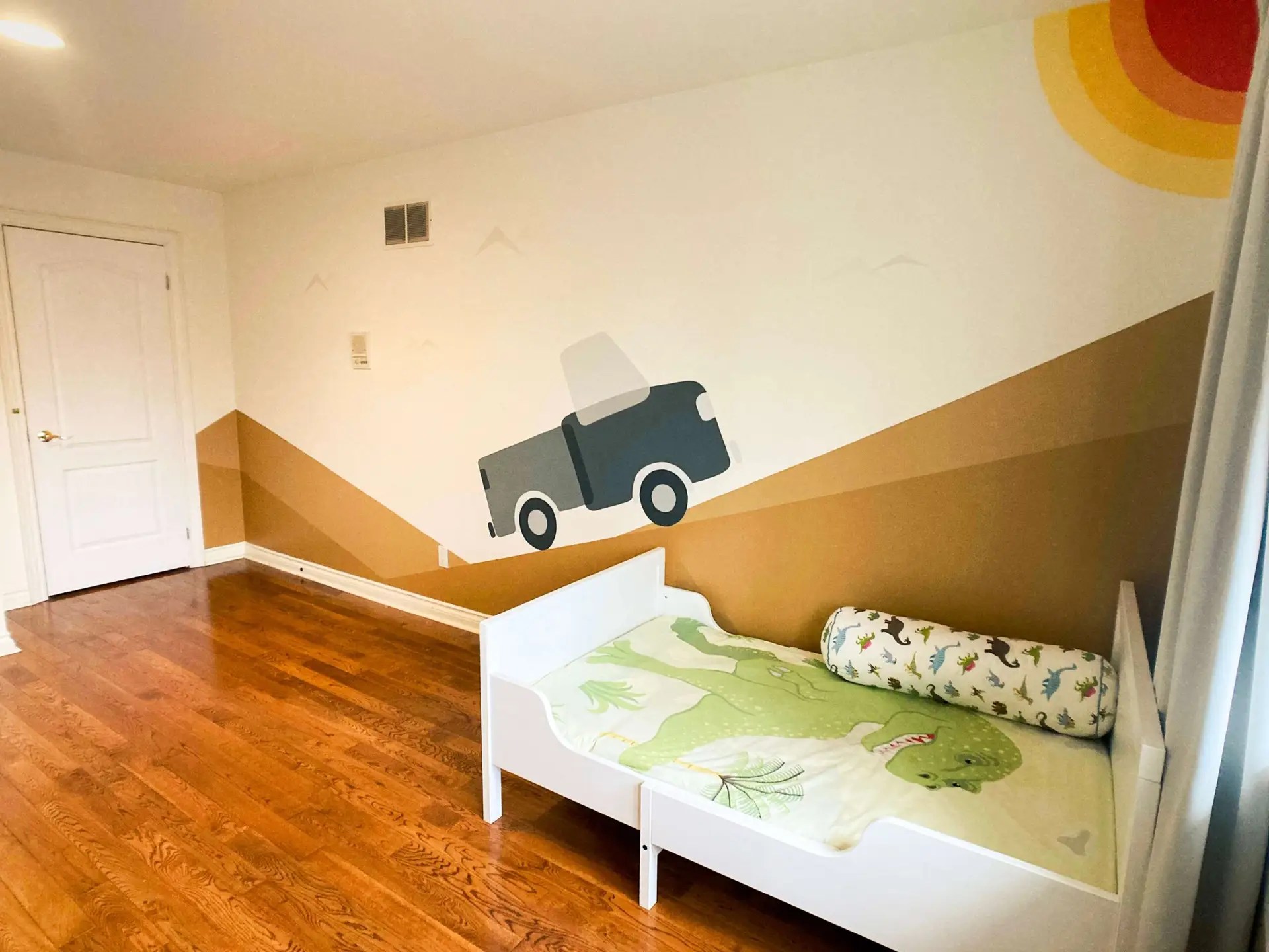 Kid's room showing a IKEA bed with dinosaur patterned bed sheets and a mural of a truck riding on the geometric background with a vibrant sun shining from the ceiling.