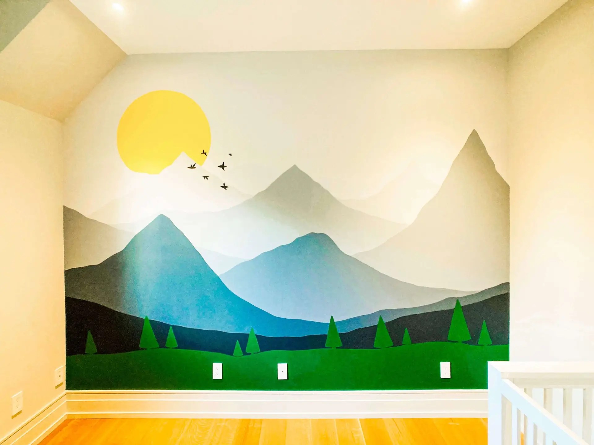 Room with a wall mural depicting a landscape full of mountains at different distances, evergreens in the foreground, a sun setting behind the mountains and a group of birds flying at the distance.
