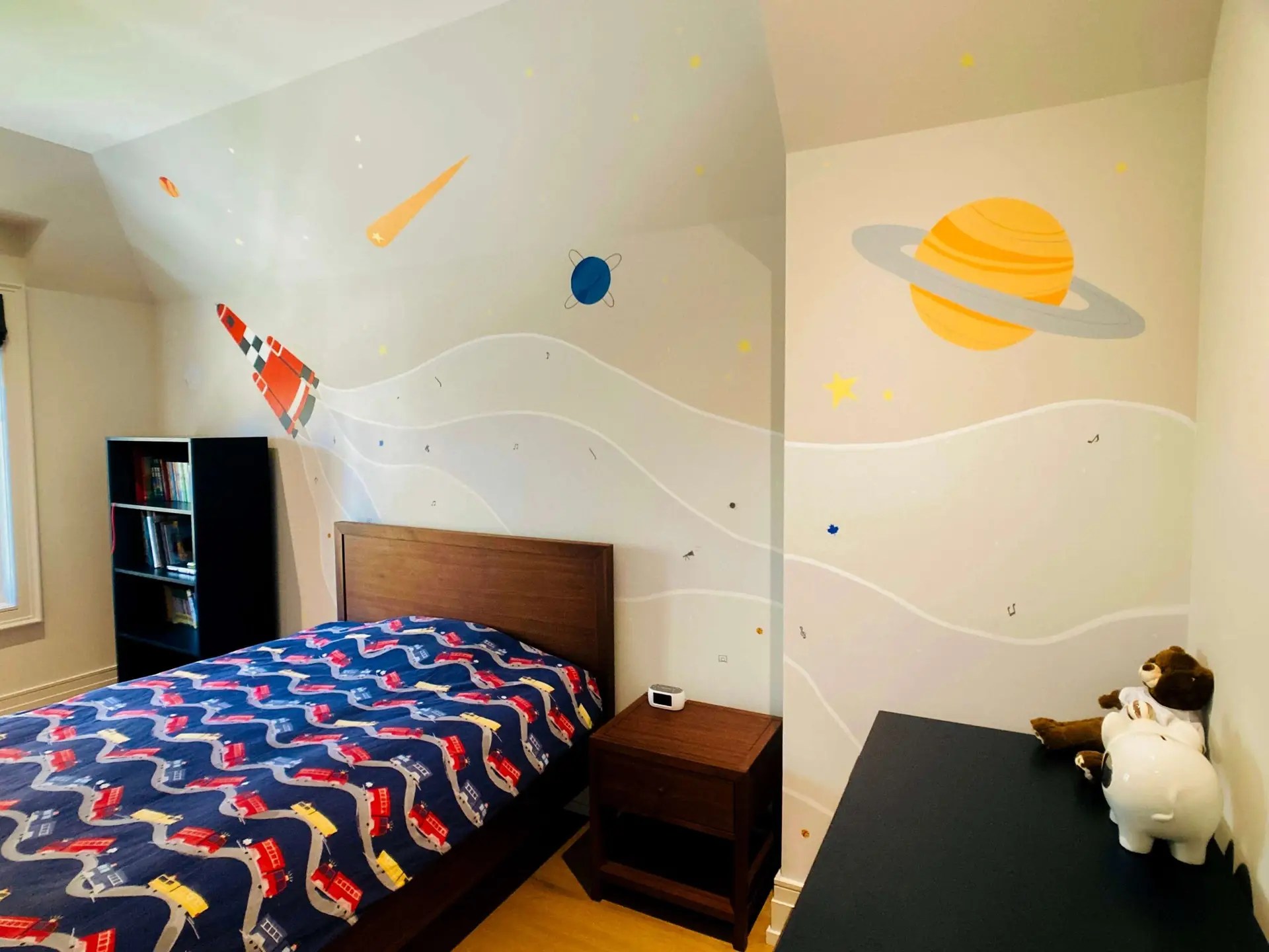Kid's bedroom with a single bed, bookcase and desk from Crate and Barrel. You can see a big out-of-space mural behind the bed with a red space rocket emitting smoke full of special particles which depict the child's interests.