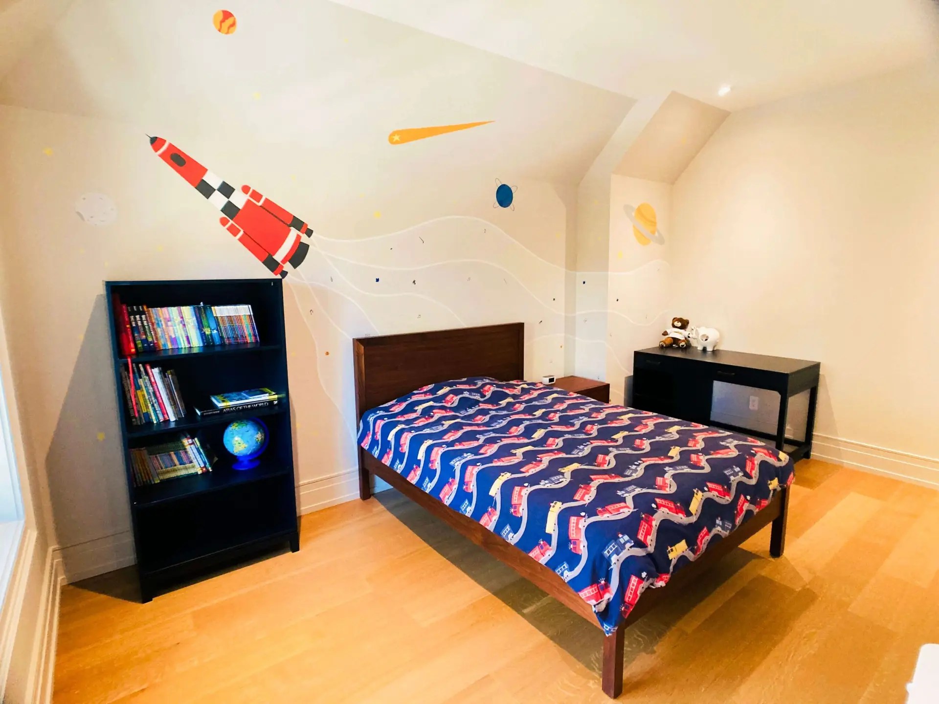 Kid's bedroom with a single bed, bookcase and desk from Crate and Barrel. You can see a big out-of-space mural behind the bed with a red space rocket emitting smoke full of special particles which depict the child's interests.