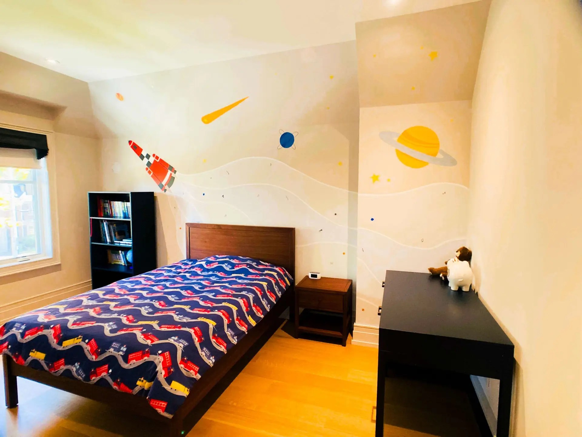 Kid's bedroom with a single bed, bookcase and desk from Crate and Barrel. You can see a big out-of-space mural behind the bed with a red space rocket emitting smoke full of special particles which depict the child's interests.