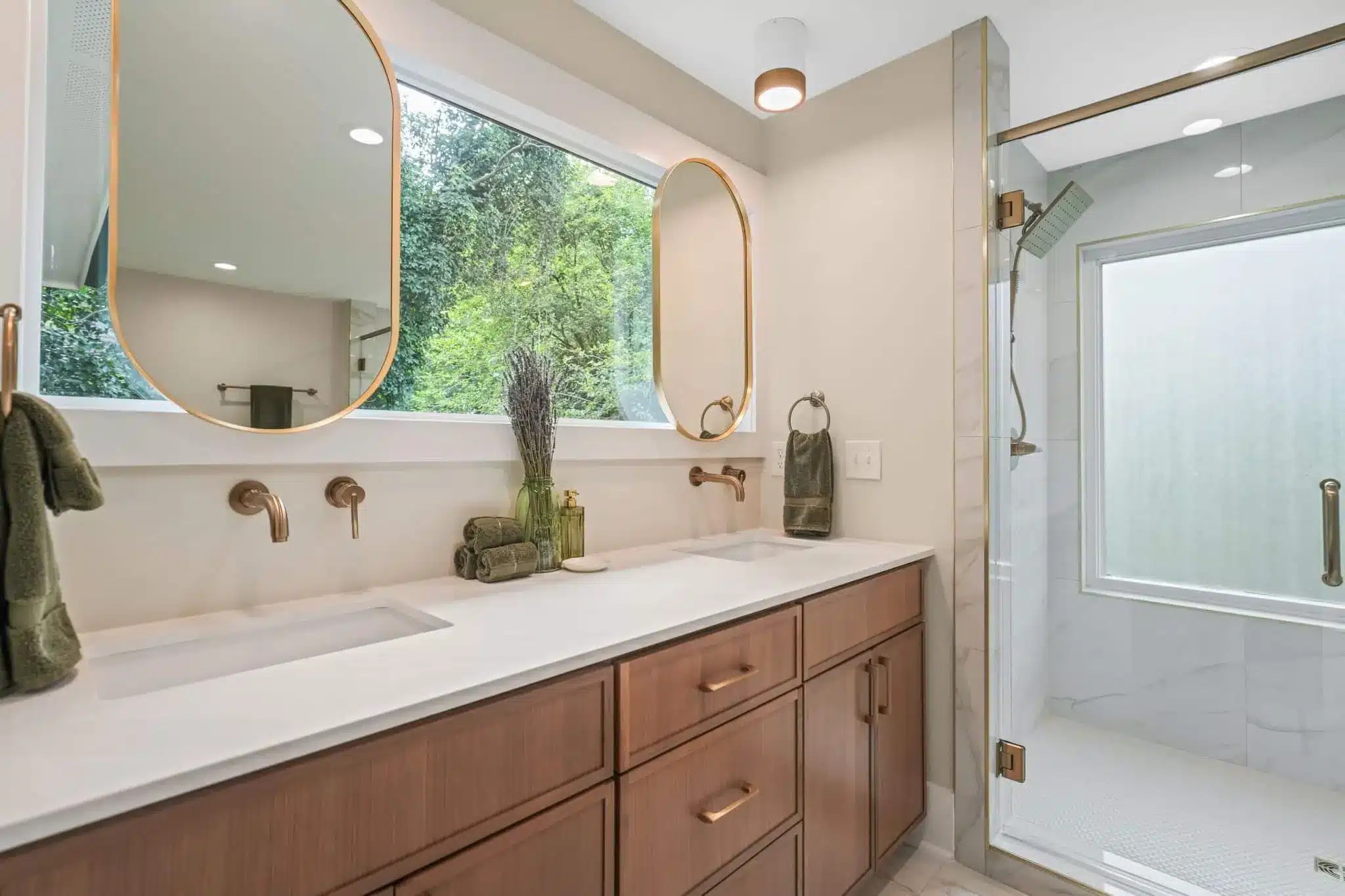 A modern luxury bathroom with a double vanity, gold-framed mirrors, white countertops, wood cabinetry, and a frameless glass shower enclosure with natural light from a large window.