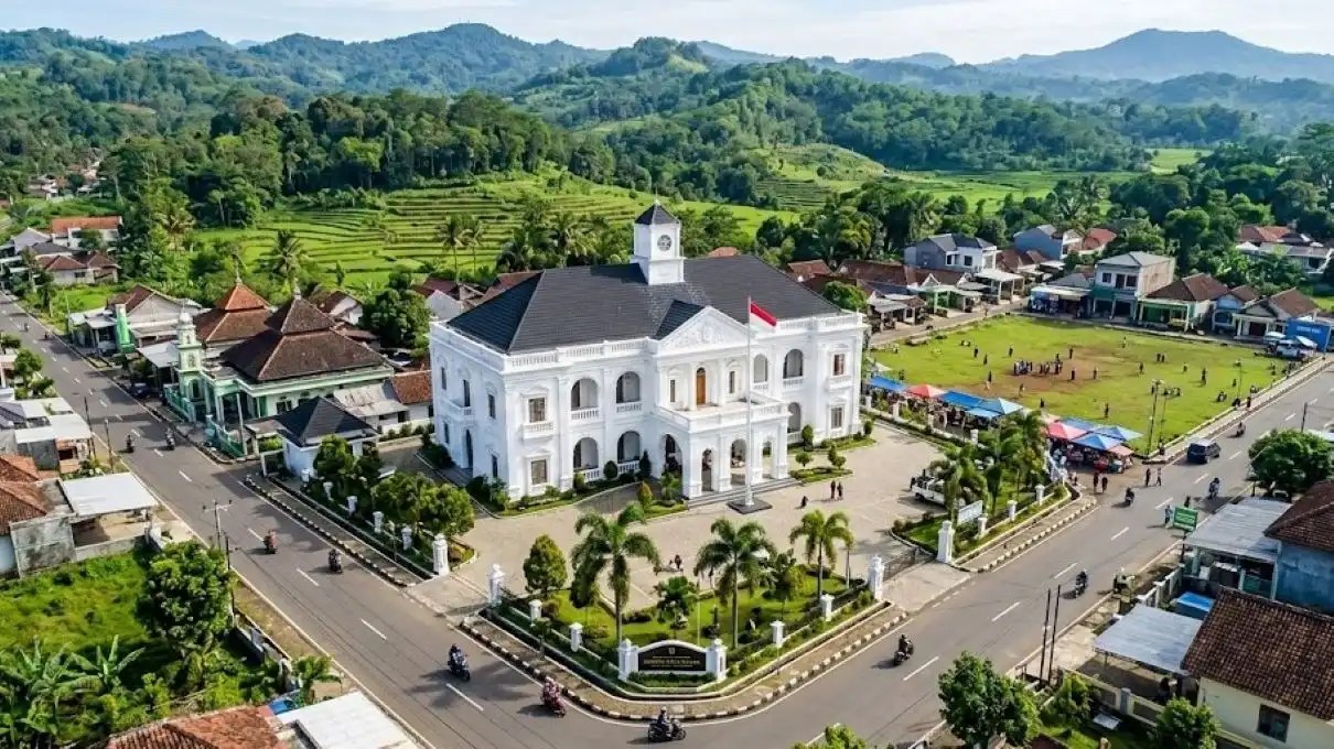 Fotografi Udara (Drone) Yang Menawan Menampilkan Kemegahan Kantor Desa Rancah Bergaya Arsitektur Klasik Eropa Berwarna Putih Bersih, Berdiri Kontras Namun Harmonis Di Tengah Lanskap Pedesaan Ciamis Yang Asri Dengan Latar Belakang Perbukitan Hijau. Di Sekitarnya, Terlihat Jalanan Rapi Dan Aktivitas Warga Lokal Yang Dinamis, Memancarkan Perpaduan Modernitas Dan Kearifan Lokal.