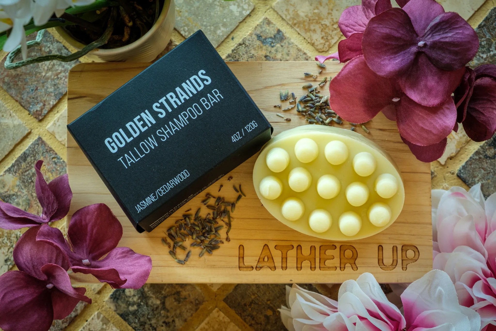 A Golden Strands Tallow Shampoo Bar and its black product box are displayed on a wooden board engraved with “LATHER UP.” The oval shampoo bar features a massage-style design with rounded nodes and is surrounded by lavender buds and vibrant floral accents, creating an elegant, spa-inspired presentation.