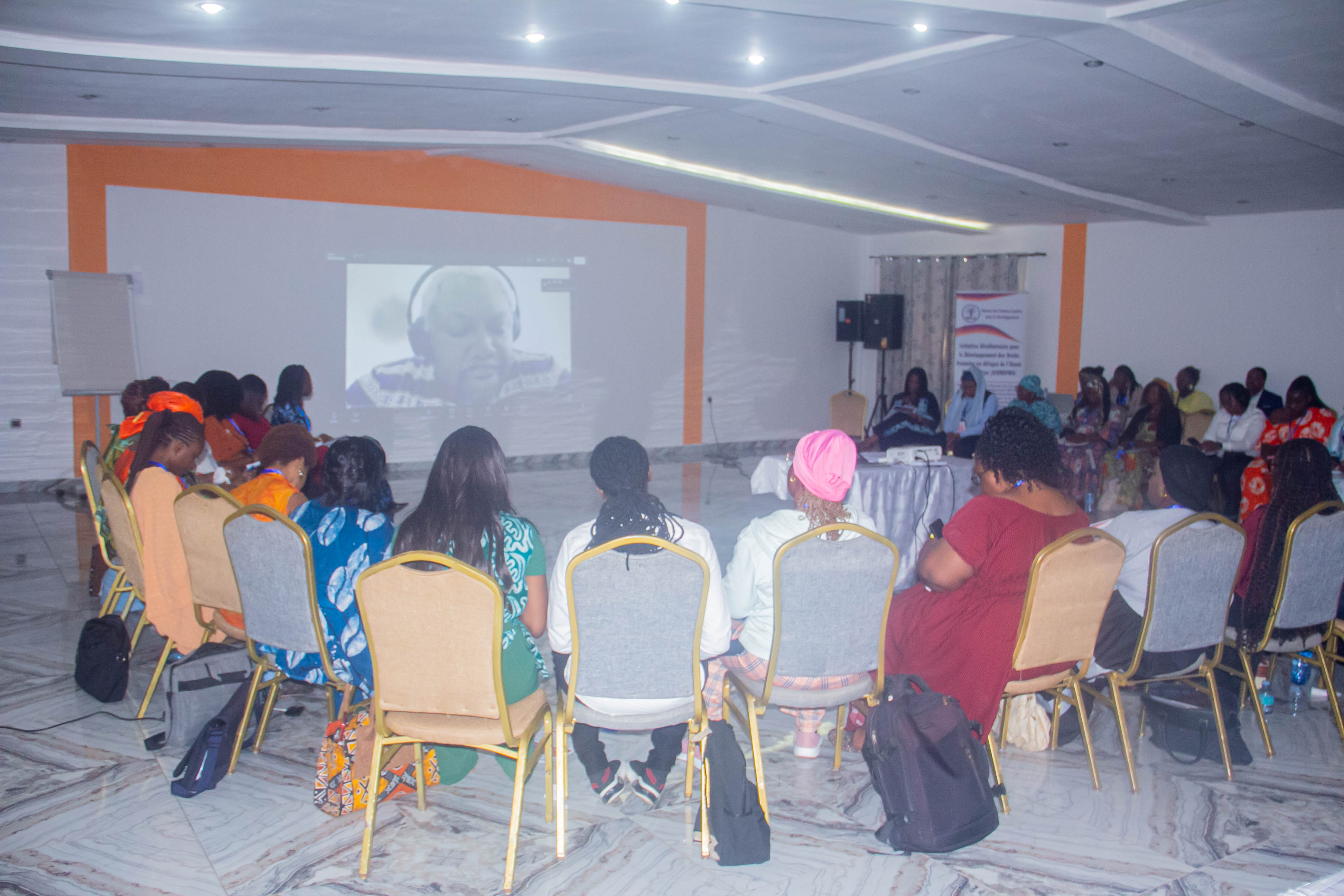 A group of people seated in a semi-circle attending a virtual meeting displayed on a screen, with a few participants visible in a conference room.
